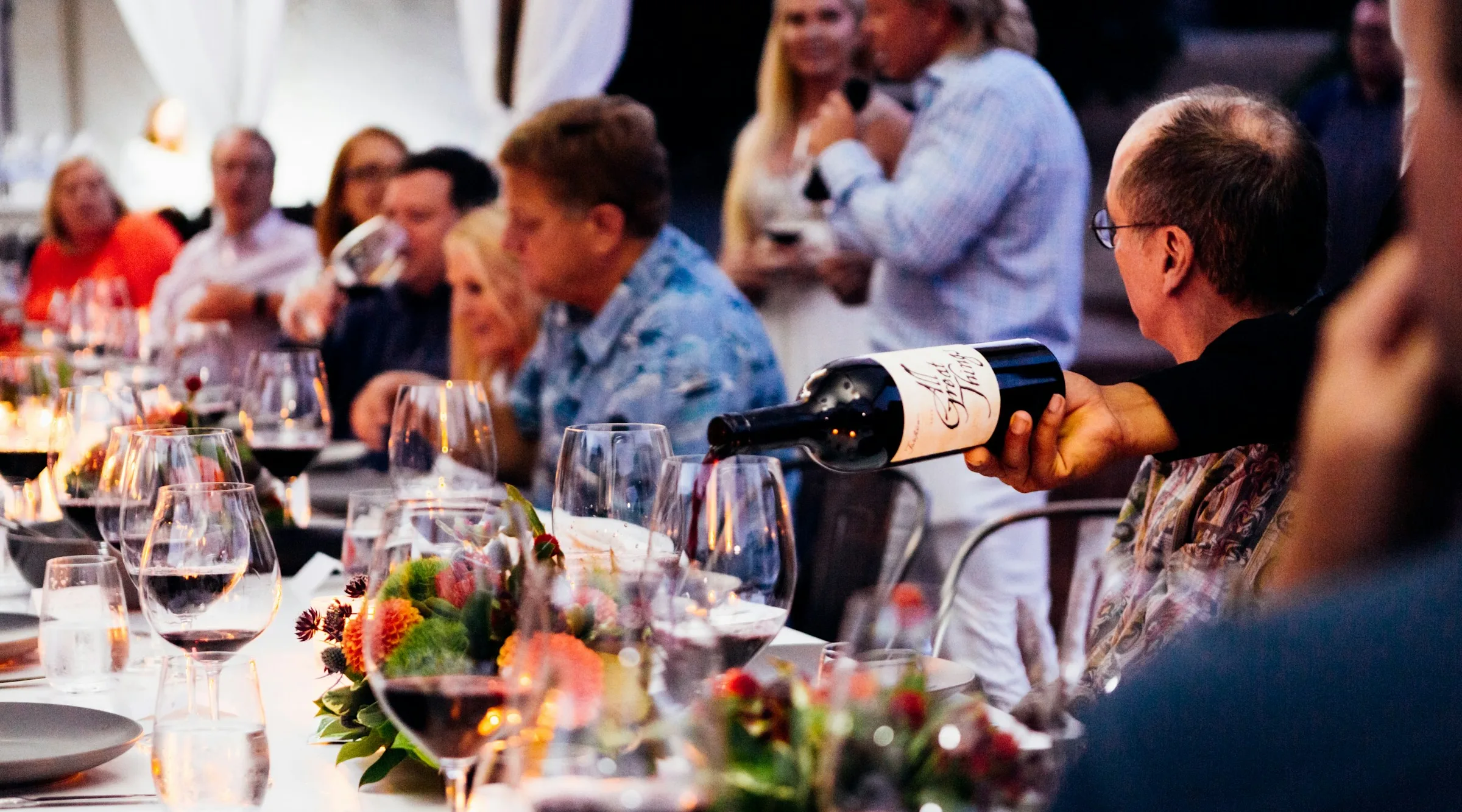 Red wine is poured along a long dinner table laid with flowers and glasses
