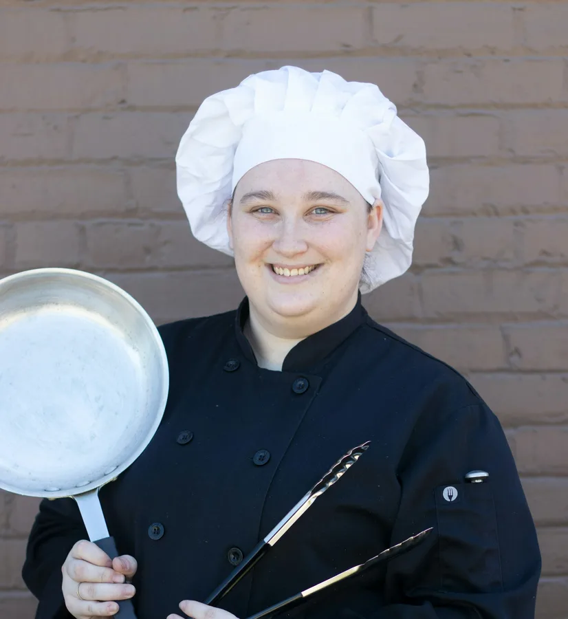 Ines Vermeulen in chef's whites, smiling, holding a frying pan and tongs
