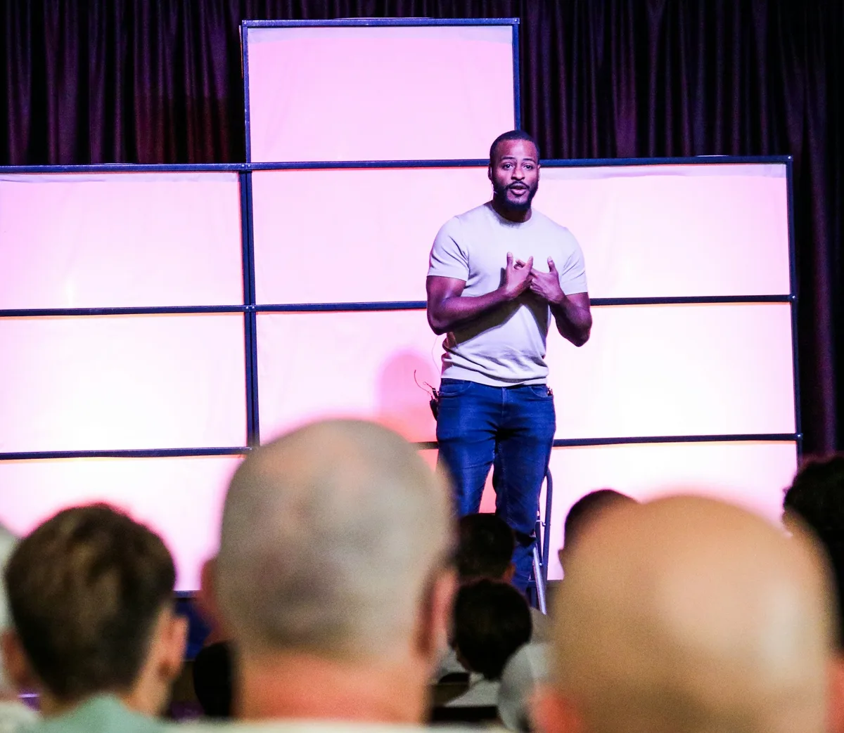 A speaker in a white T-shirt talks from a lit stage to a packed room
