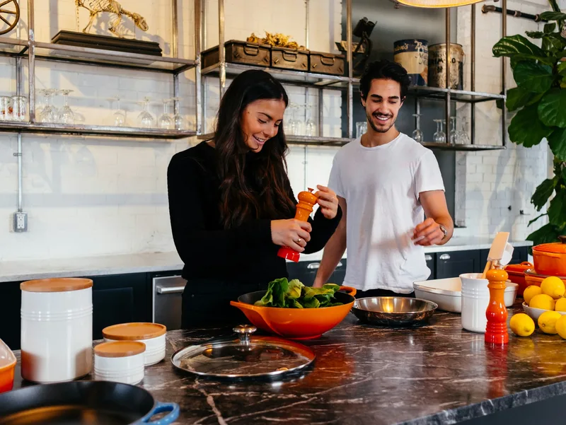 Two cooks season a bowl of greens beside a bowl of lemons