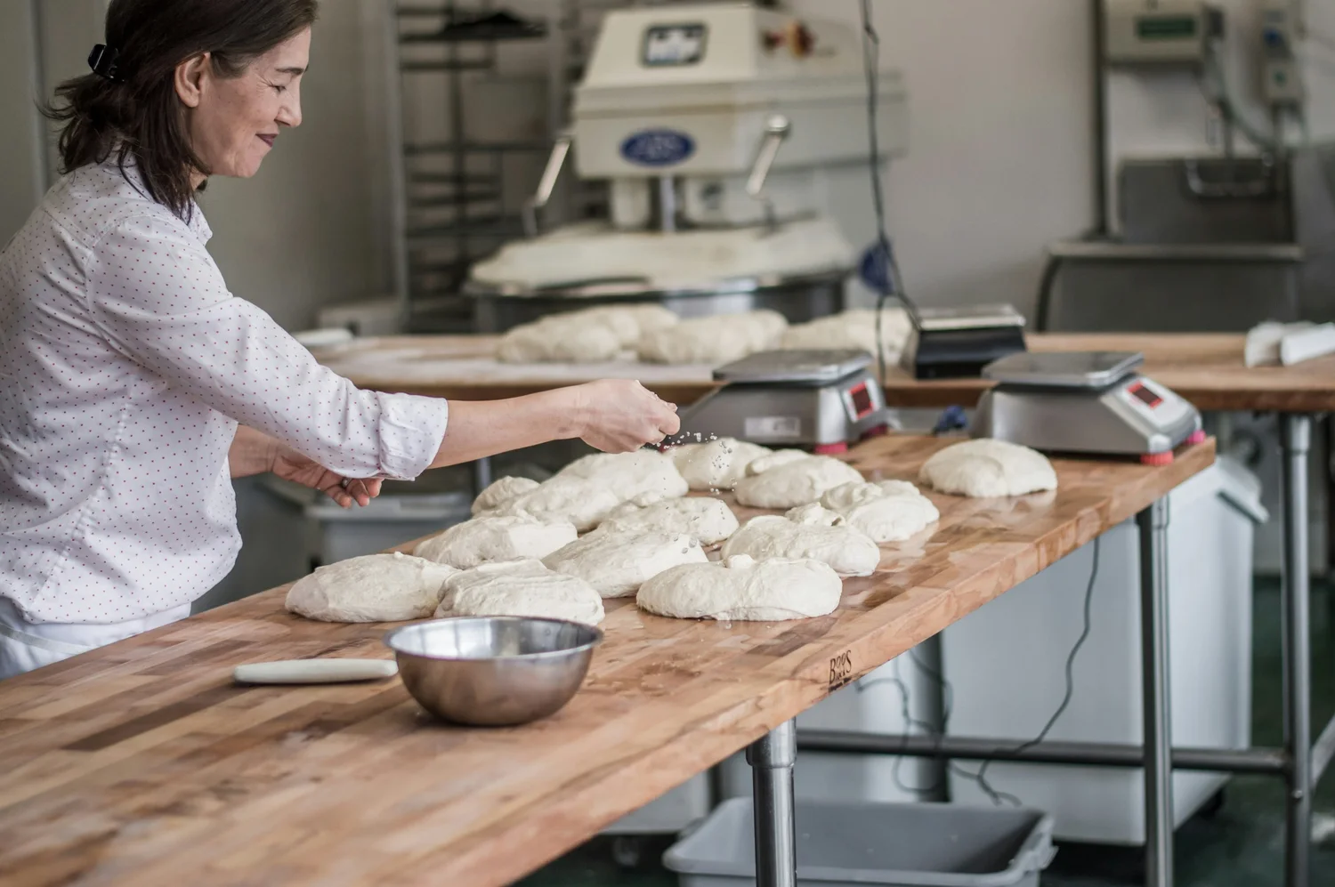 A baker smiles as she divides dough into rounds along a long wooden bench