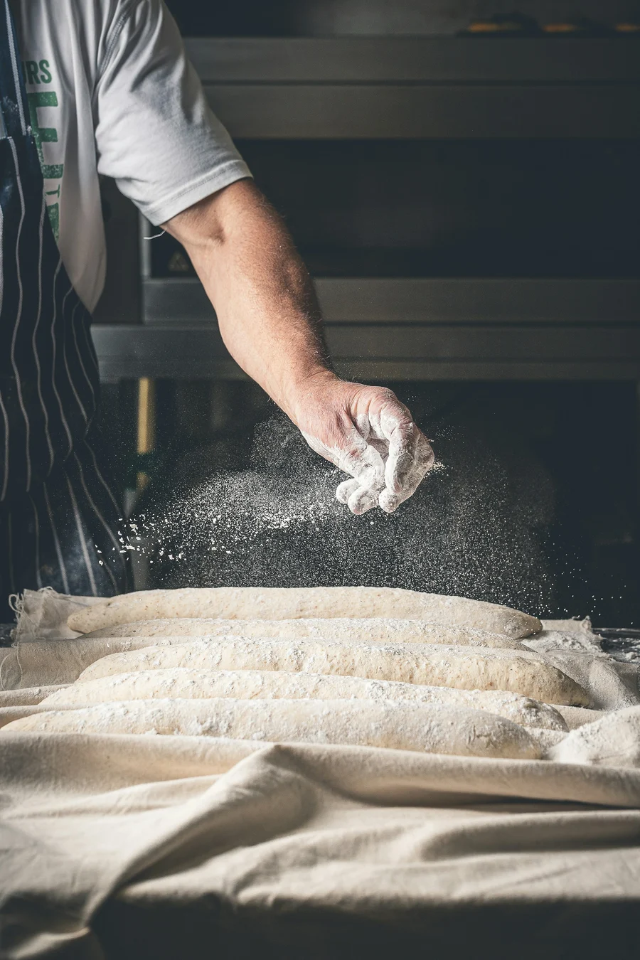 A baker scatters flour over a row of proofing loaves on linen