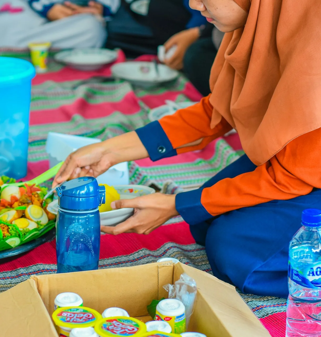 A volunteer in an orange hijab serves rice and salad onto plates laid out on a floor mat