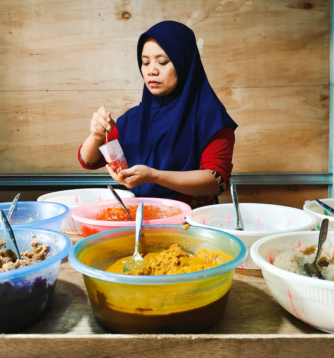 A kitchen volunteer spoons sambal into bags beside bowls of curry and rice prepared for iftar