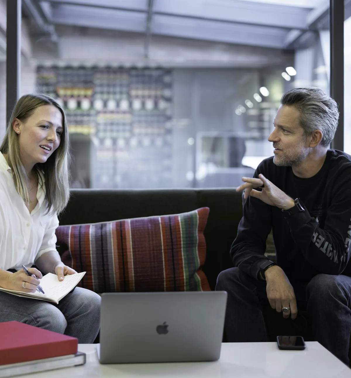 An Ashby & Lane bookkeeper with a notebook talking a studio founder through his month on a sofa beside a laptop