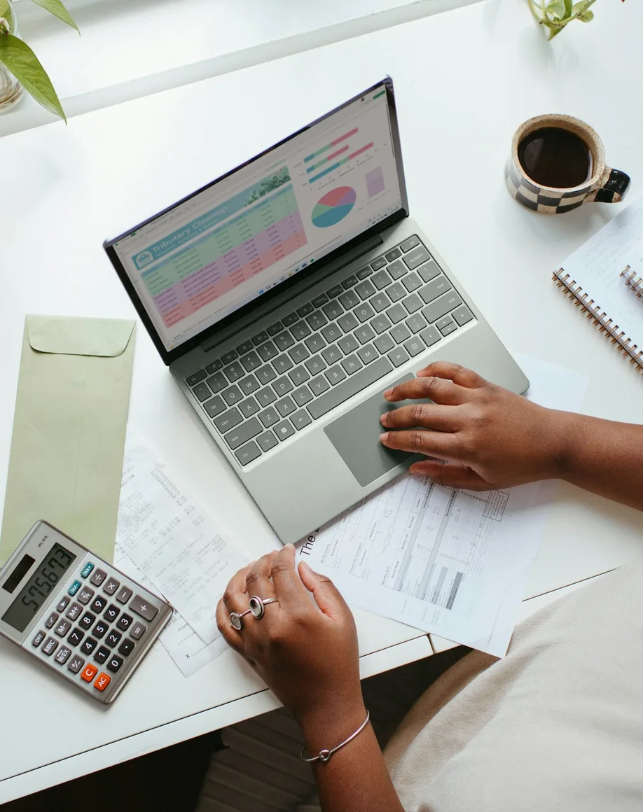 Hands on a laptop spreadsheet beside receipts, a calculator and a notebook on a white desk