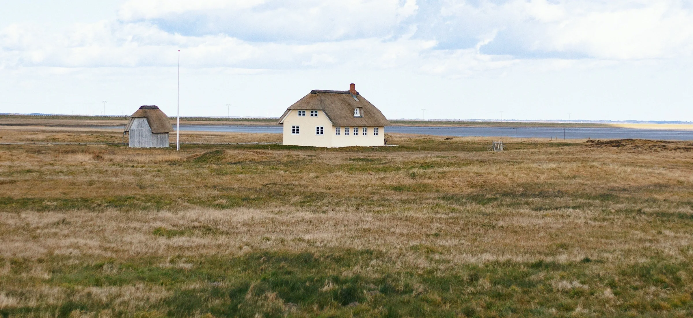 Two low houses on open grassland under a wide pale sky in Jutland