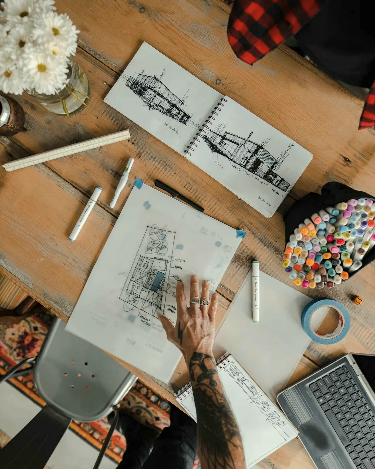Top-down view of a wooden drawing table with a sketchbook of house drawings, markers and a hand on a plan