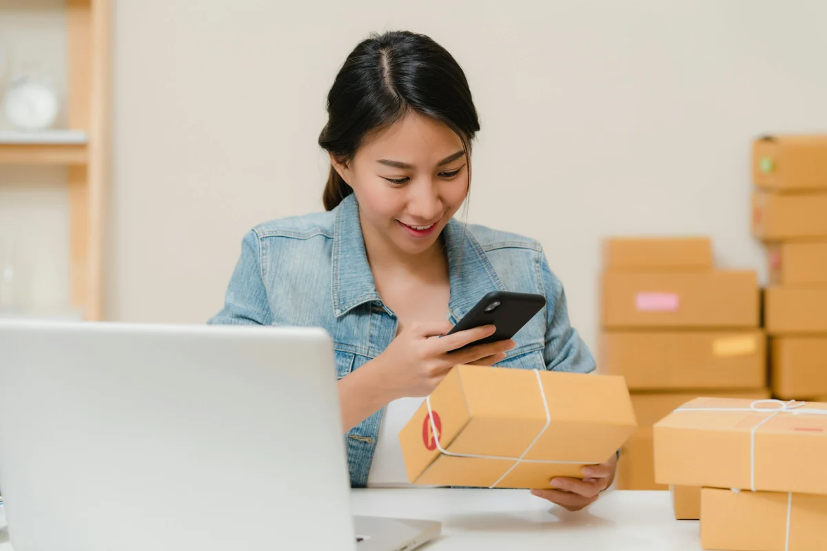 A shop owner checks an order on her phone beside a stack of packed parcels