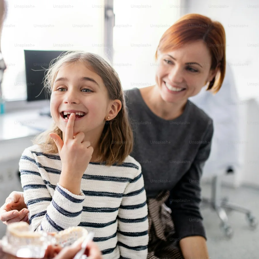 A girl in a striped top laughs and points at her tooth next to her mom
