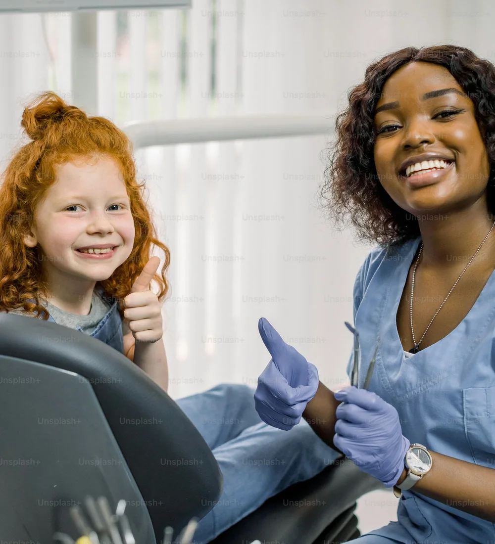 A red-haired girl in the dental chair gives a thumbs up next to her smiling dentist