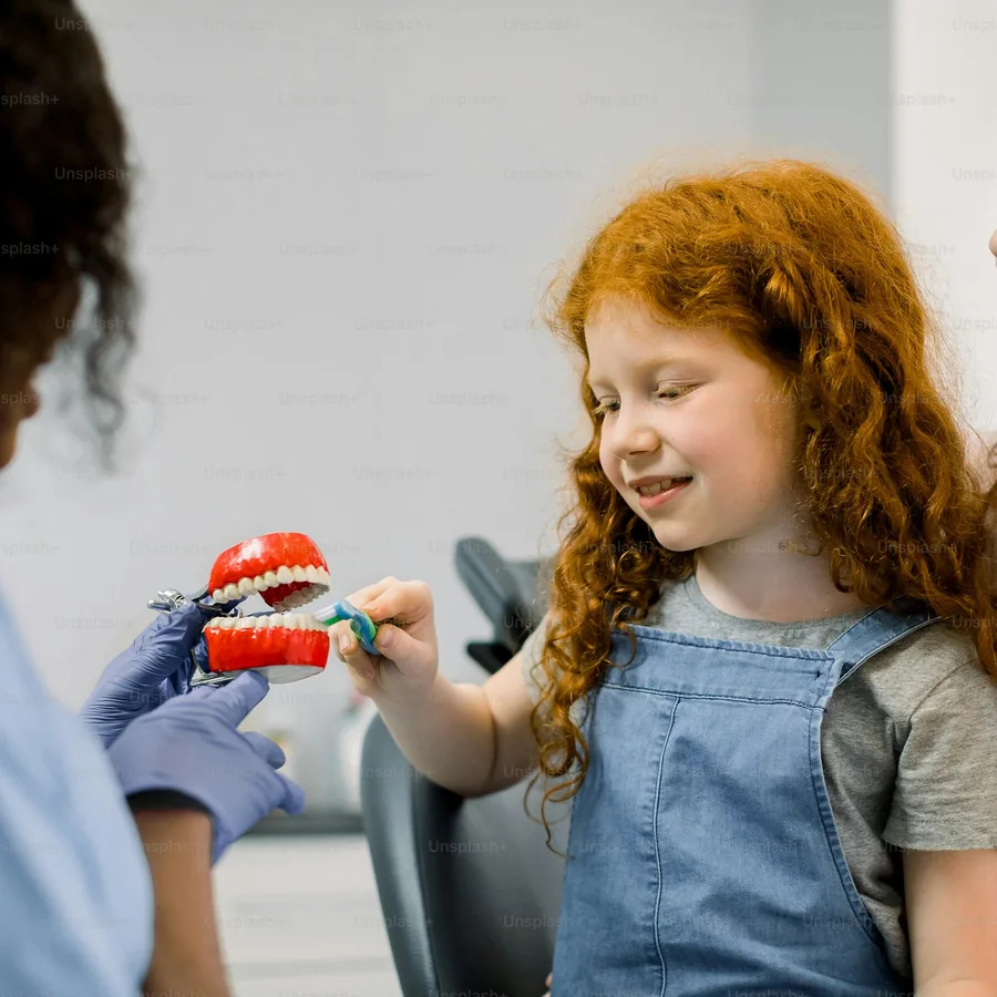 A red-haired girl smiles as she brushes a big toy tooth model