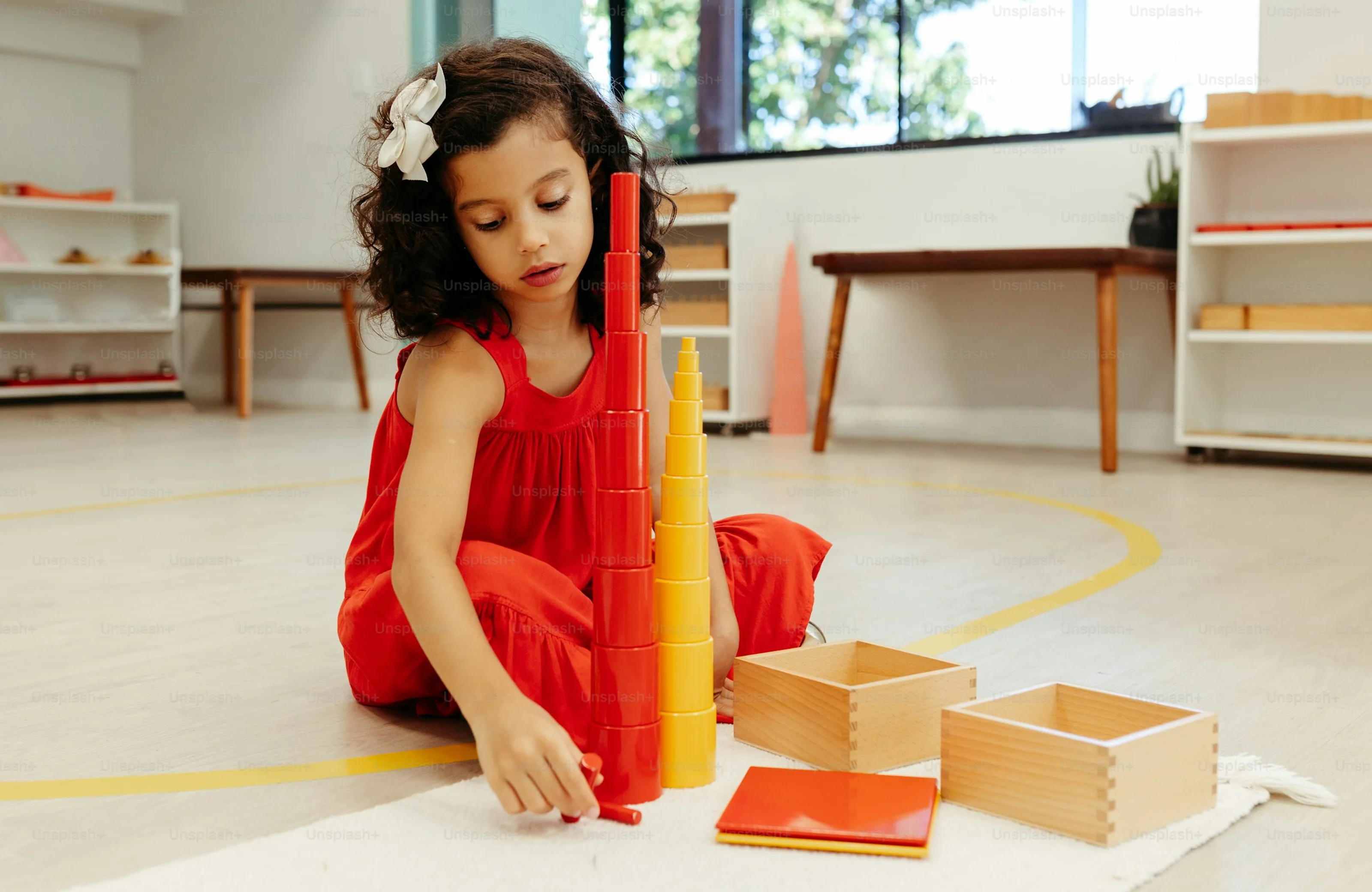 A girl in a red dress builds a pink tower on a mat in the classroom