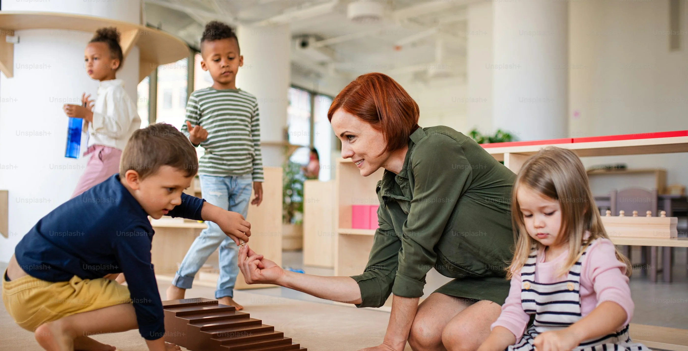 A teacher kneels on the classroom floor helping children with wooden Montessori materials