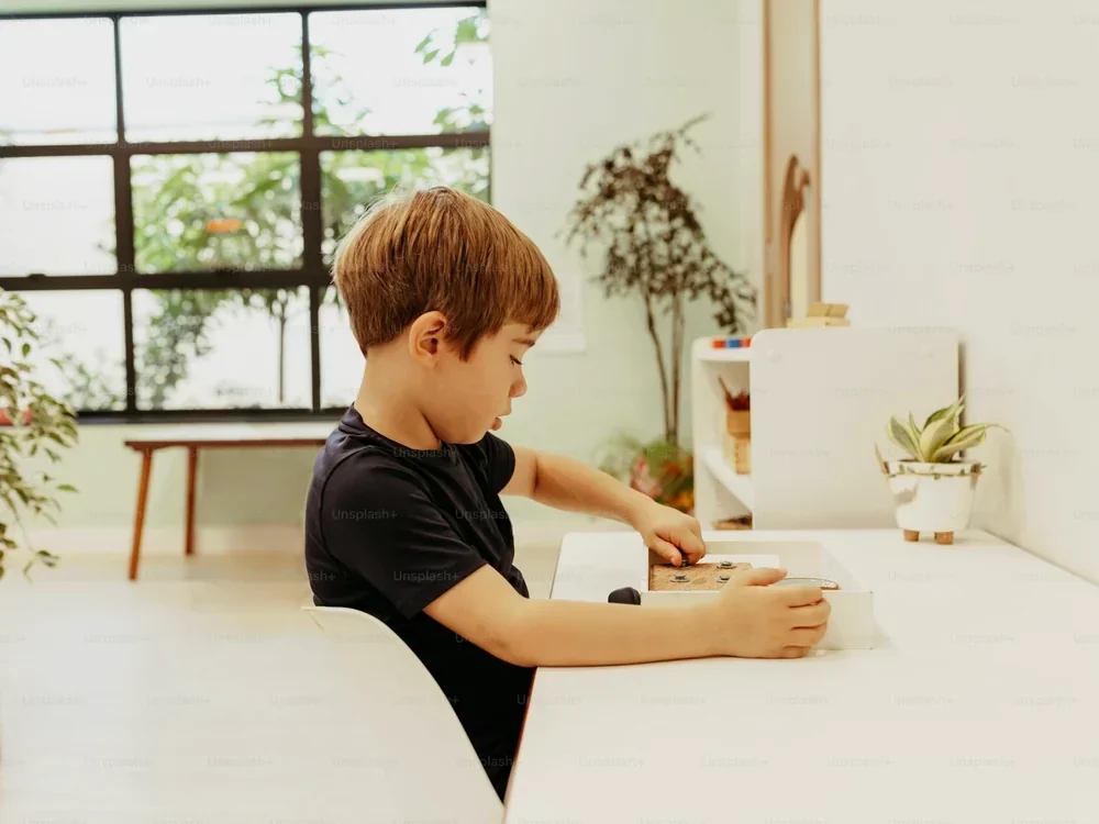 A boy works with a wooden tray at a low white table by the window