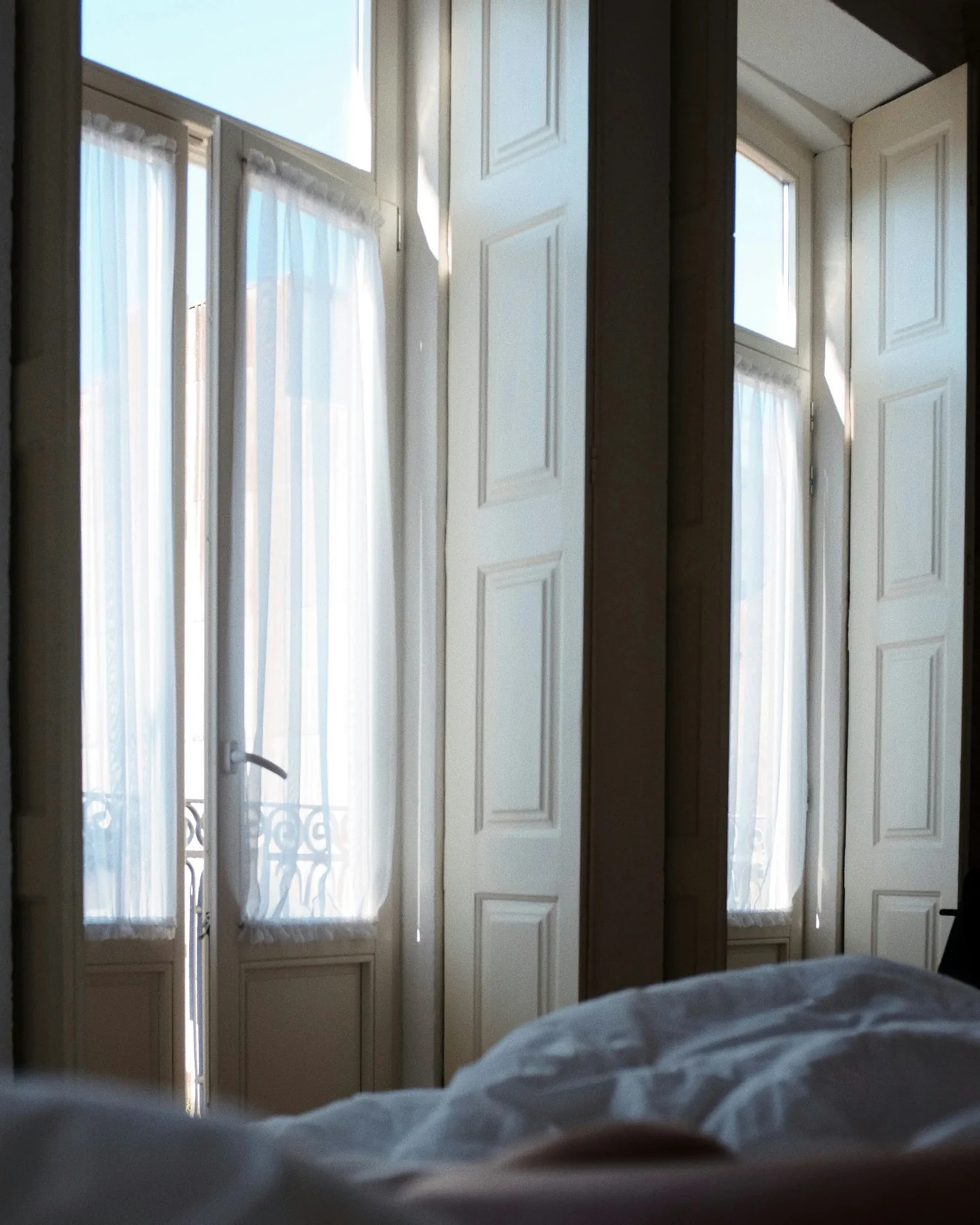 A Casa Lumen room: two tall windows with their shutters folded back, sheer curtains and white linen in morning sun
