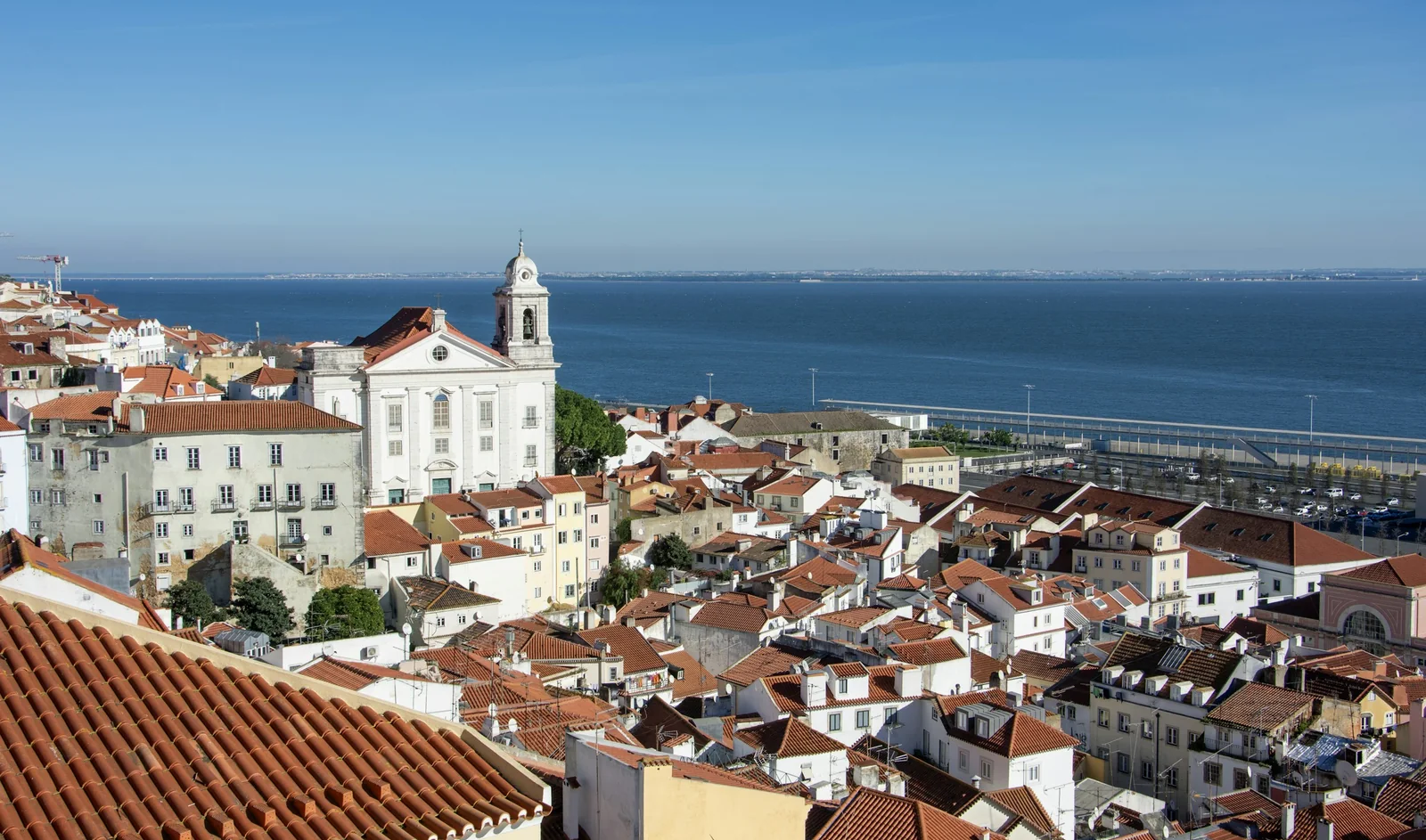 Alfama from the roof of Casa Lumen: white houses, terracotta roofs and the church of Santo Estêvão above the Tejo in morning sun