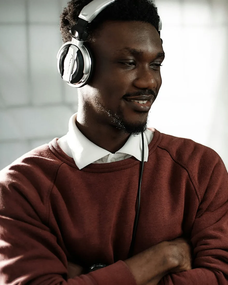 Kofi Mensah in headphones beside his microphone, smiling at an edit on screen