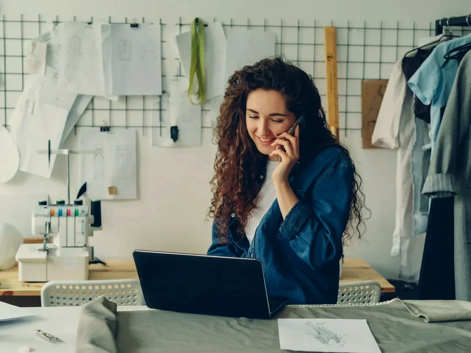 A fashion-studio owner on the phone at her laptop among clothes rails and pattern sketches