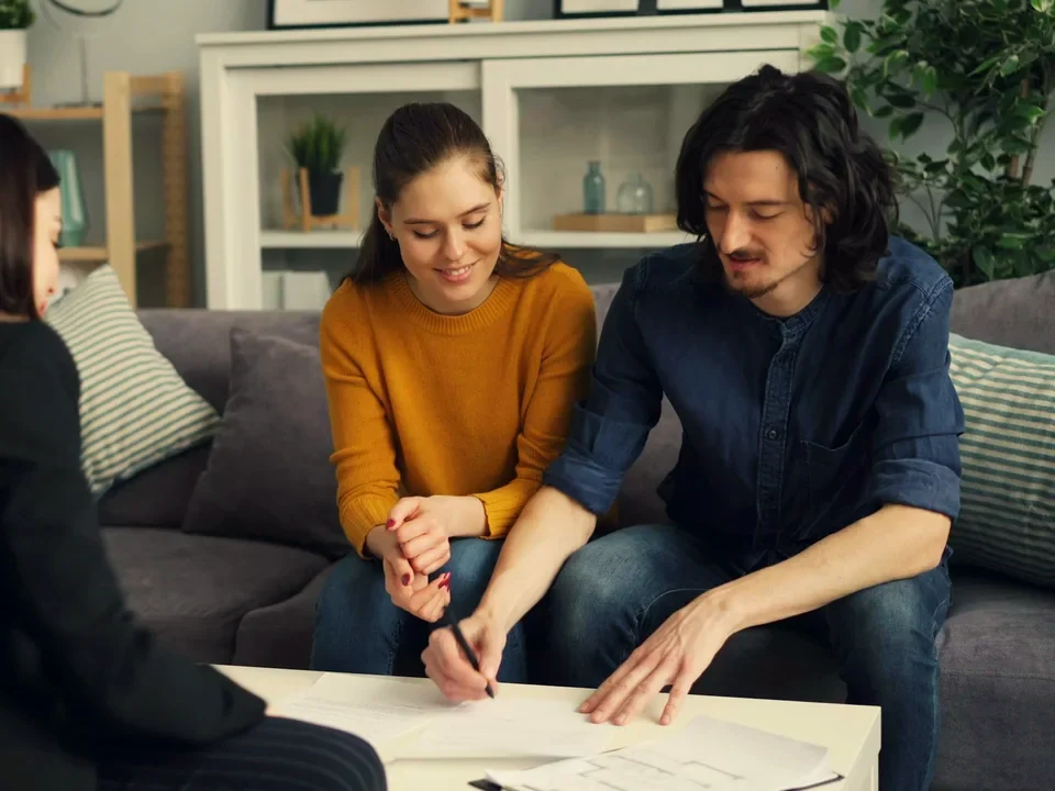 An adviser with a young couple on their sofa, the man signing papers on the coffee table