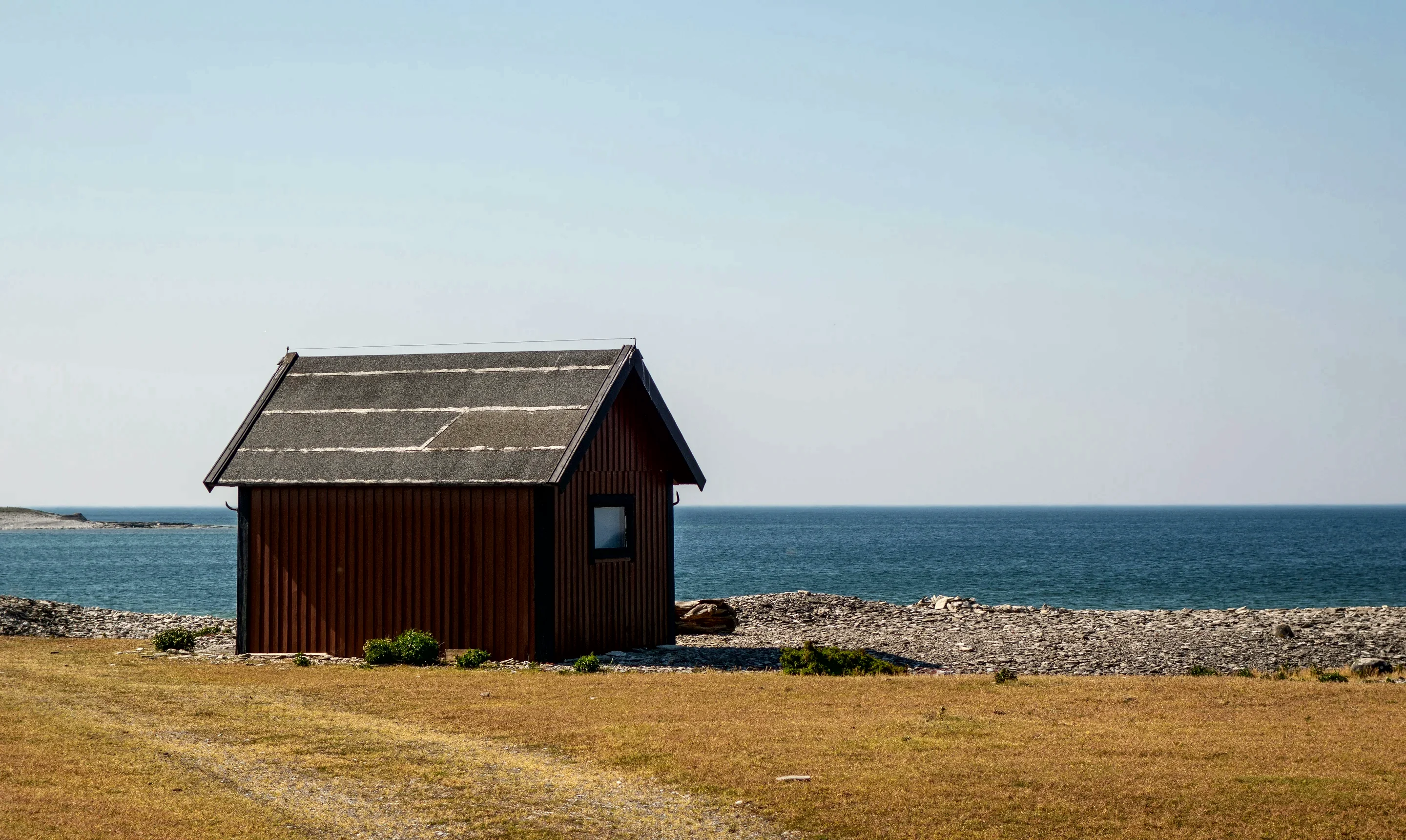 A dark timber sauna cabin on a pebble shore with the sea and a clear sky behind it