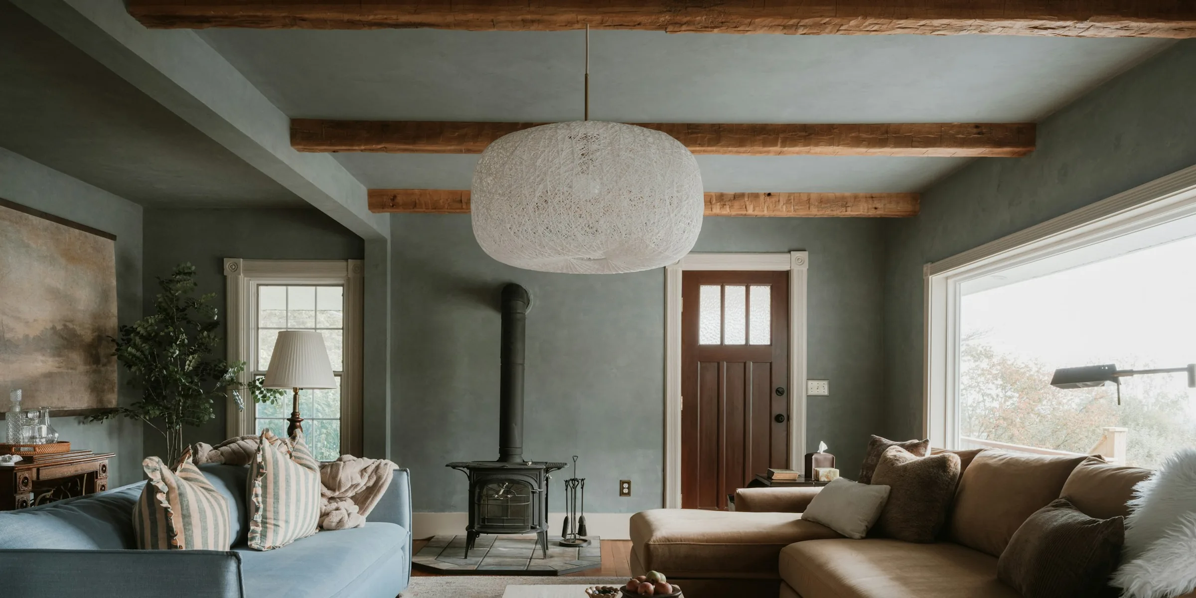 A daylight living room with timber ceiling beams, a wood stove, a tan sofa and a large garden window