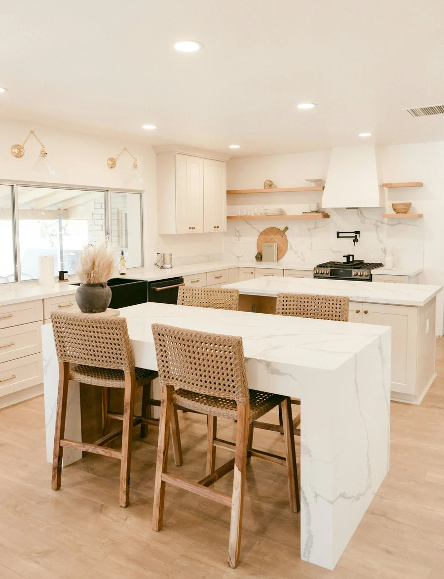 A white kitchen with a marble island and woven timber stools on a pale oak floor
