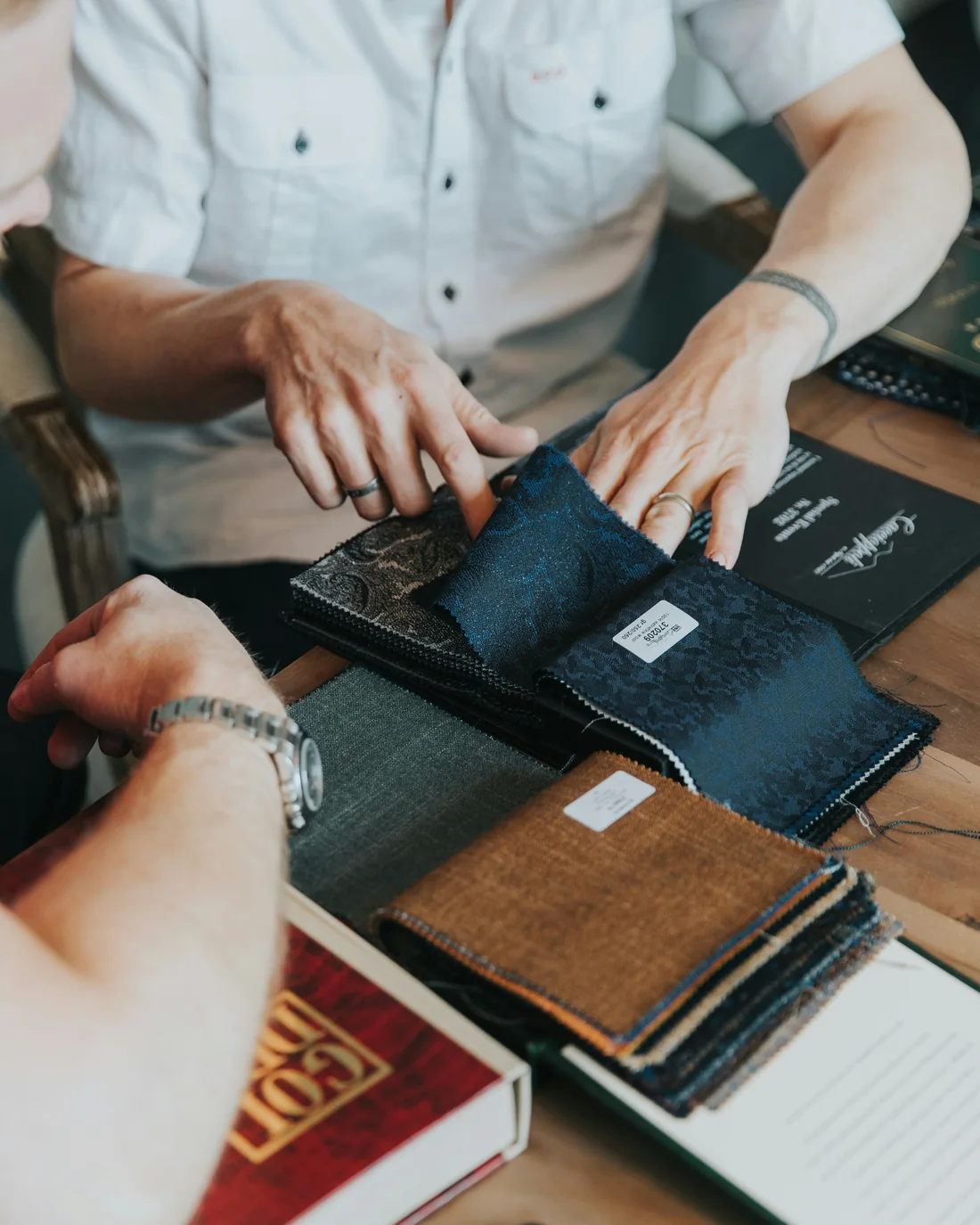 A designer's hands open a book of dark fabric samples on a timber desk