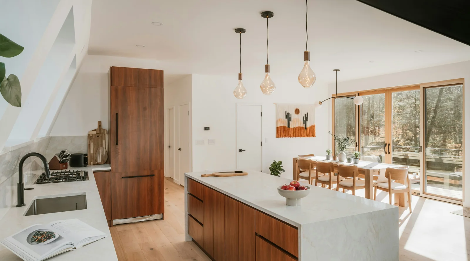 A long kitchen with a spotted gum island, a stone benchtop, three glass pendants and a dining table by the garden windows