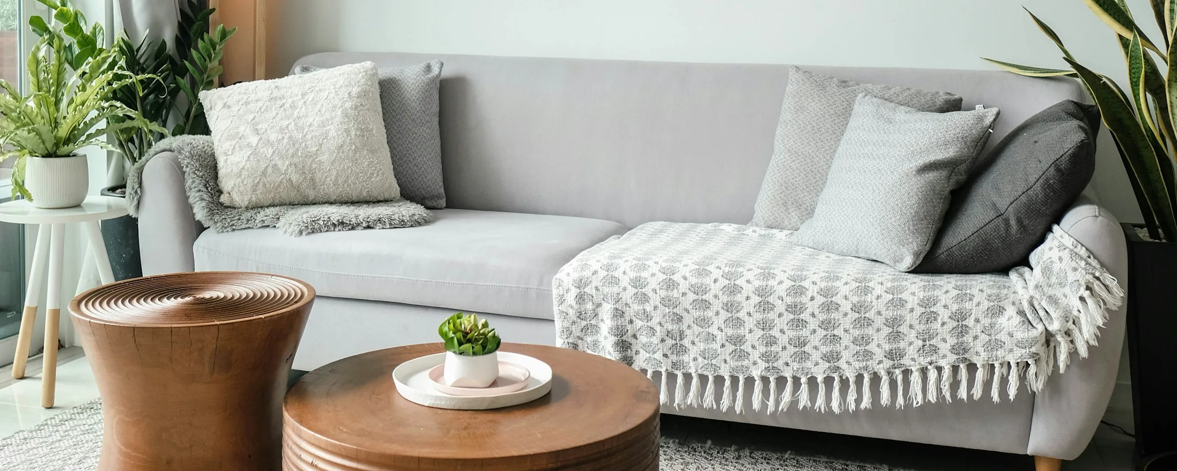 A finished living room in soft daylight: a pale linen sofa with a knitted throw, two turned timber side tables and plants by the window