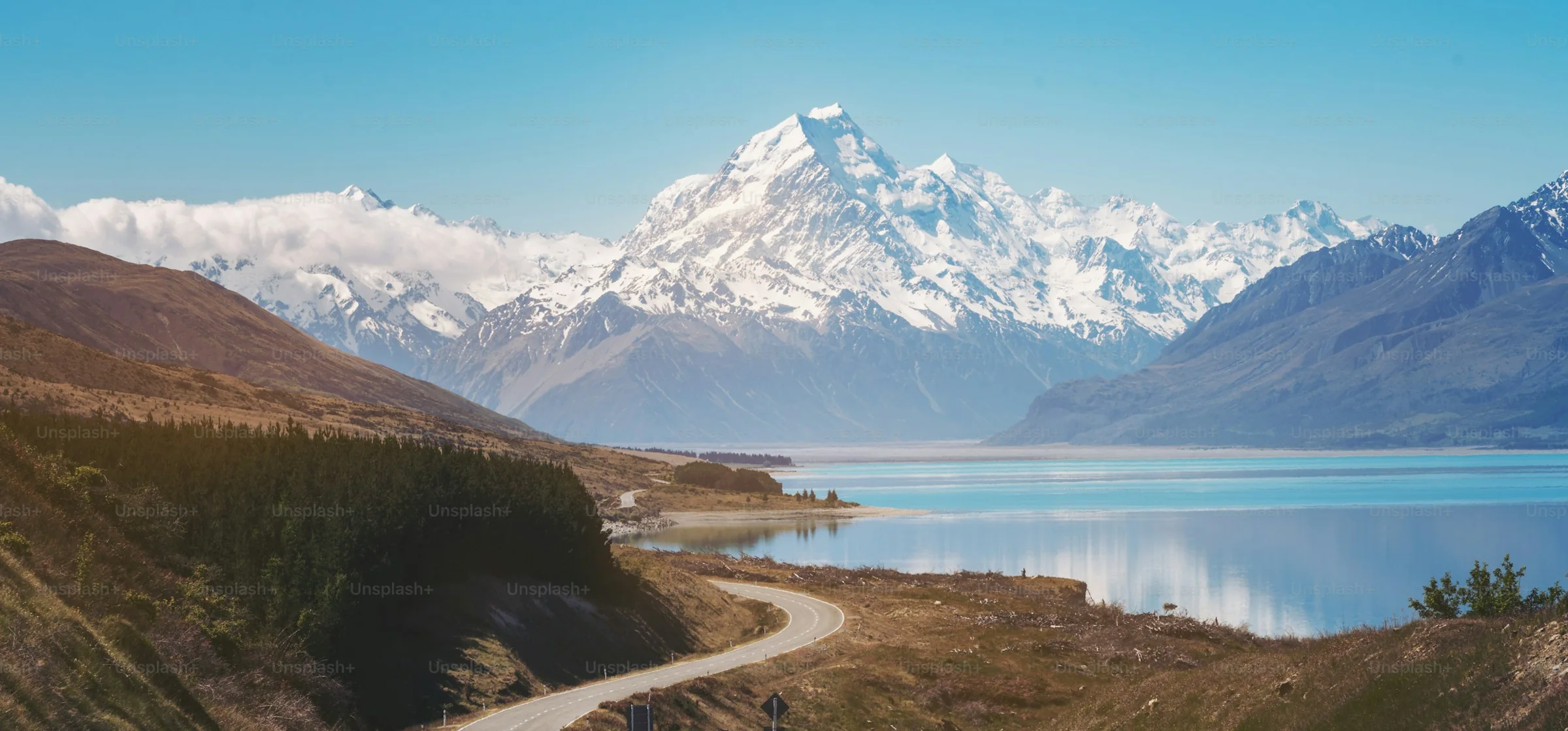 A winding road running toward a turquoise lake and a snow-capped peak under a clear blue sky