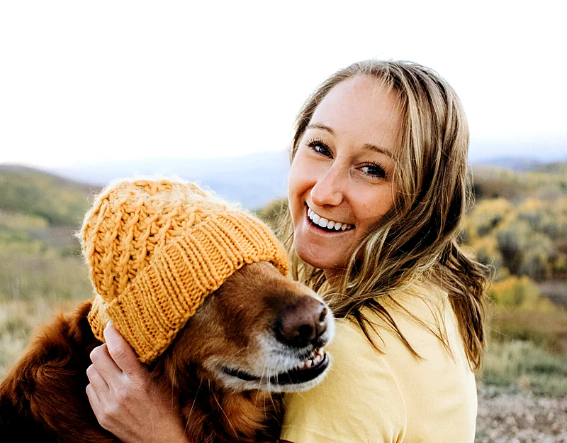 A laughing woman cuddling her dog in a knitted hat on a hillside walk