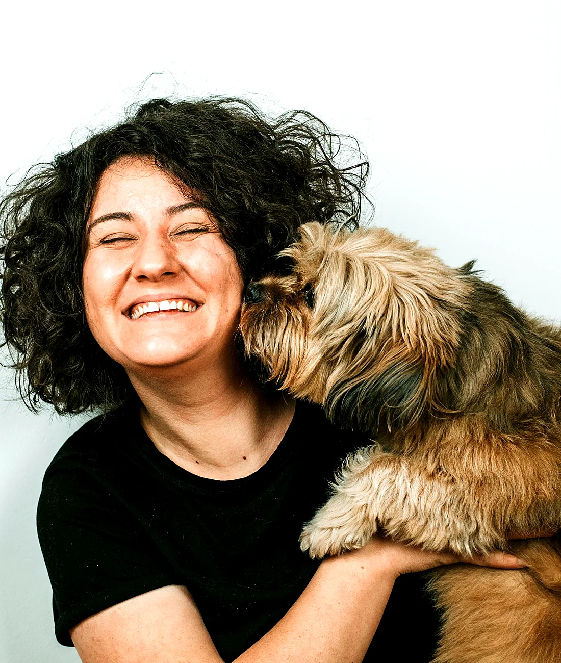 A laughing woman being nuzzled by her shaggy terrier against a bright white wall